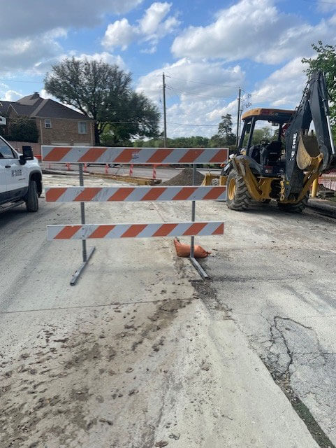Road construction blocking through traffic with excavator and open trench across the street near the community entrance.